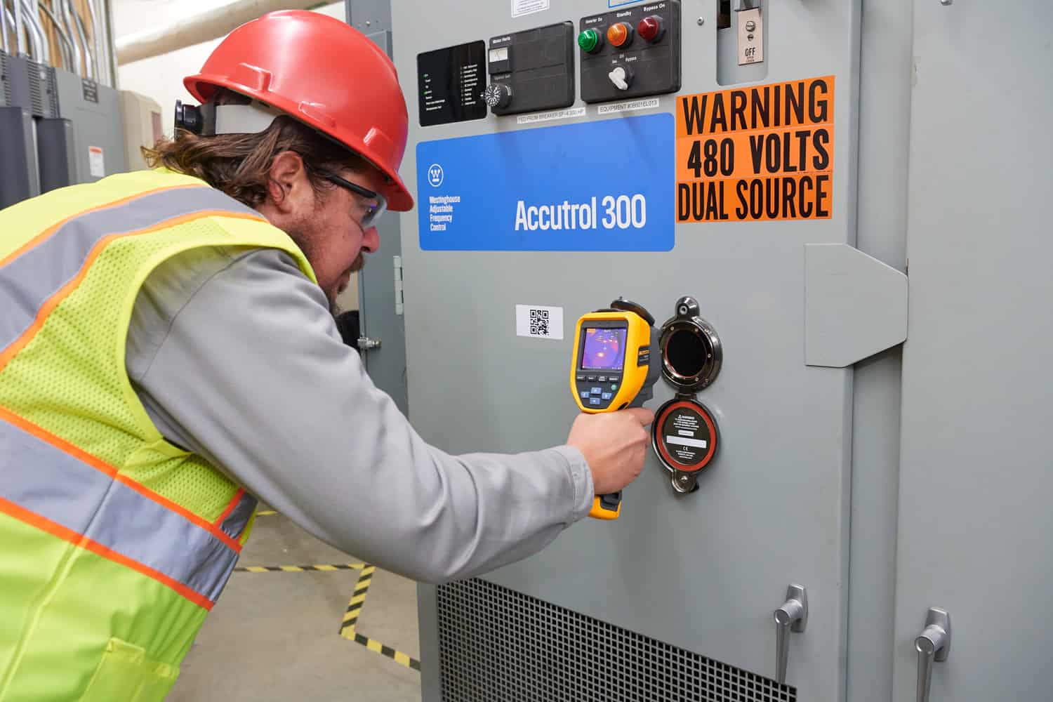 วิศวกรสวมหมวกนิรภัยสีแดงและเสื้อกั๊กสะท้อนแสง กำลังใช้กล้องถ่ายภาพความร้อน Fluke ตรวจสอบจุดเชื่อมต่อไฟฟ้าภายในตู้ควบคุม Accutrol 300 เพื่อวิเคราะห์ความร้อนสะสมในระบบไฟฟ้า 480 โวลต์