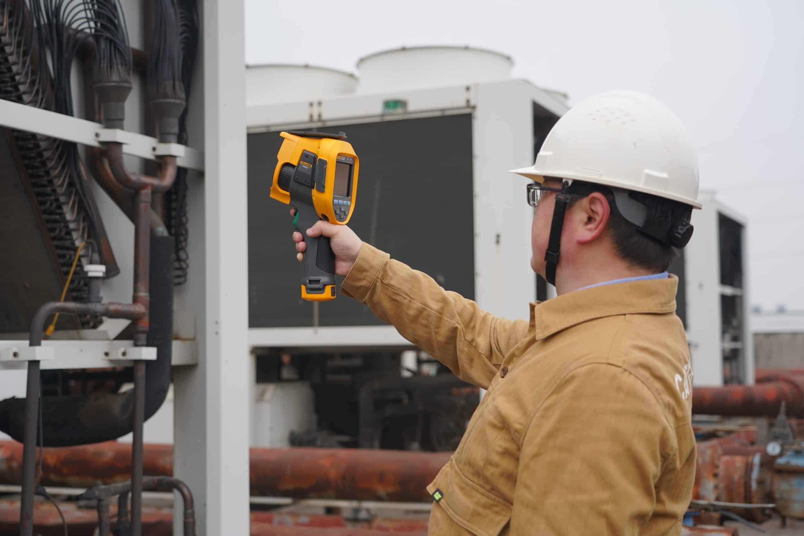 วิศวกรซ่อมบำรุงสวมหมวกนิรภัยสีขาว กำลังใช้กล้องถ่ายภาพความร้อน Fluke ตรวจสอบอุณหภูมิของระบบคูลลิ่งทาวเวอร์ (Cooling Tower) และท่อส่งน้ำบนดาดฟ้าโรงงาน