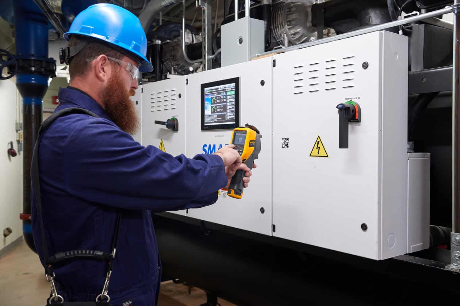 วิศวกรสวมหมวกนิรภัยสีน้ำเงิน กำลังใช้กล้องถ่ายภาพความร้อน Fluke ตรวจสอบตู้ควบคุมไฟฟ้า Smart Panel ที่มีหน้าจอแสดงผลและ QR Code โดยใช้ฟีเจอร์ Asset Tagging เพื่อจัดเก็บข้อมูลภาพถ่ายความร้อนอย่างเป็นระบบ