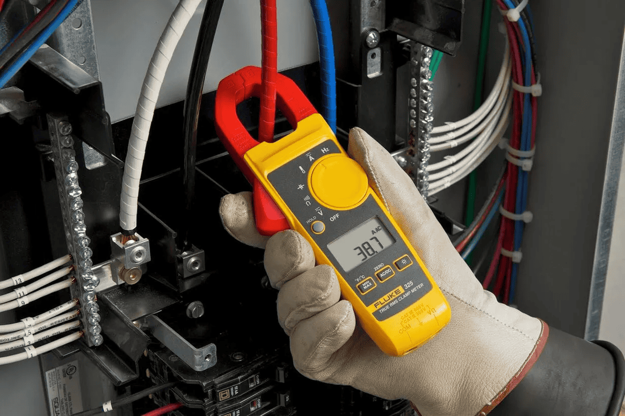 A hand wearing a protective safety glove holding the Fluke 325 True-RMS Clamp Meter measuring AC current inside an industrial electrical control panel